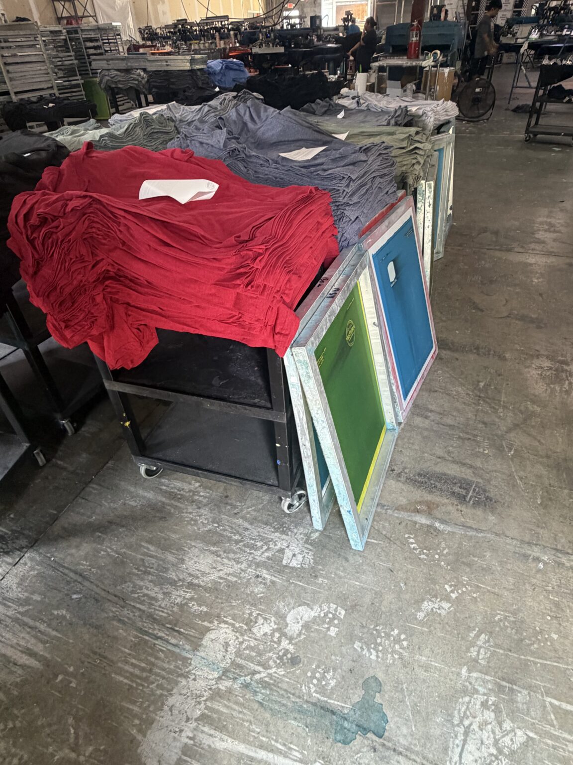 Cart stacked with folded red and heather shirts next to screen printing frames on the 5Boys production floor